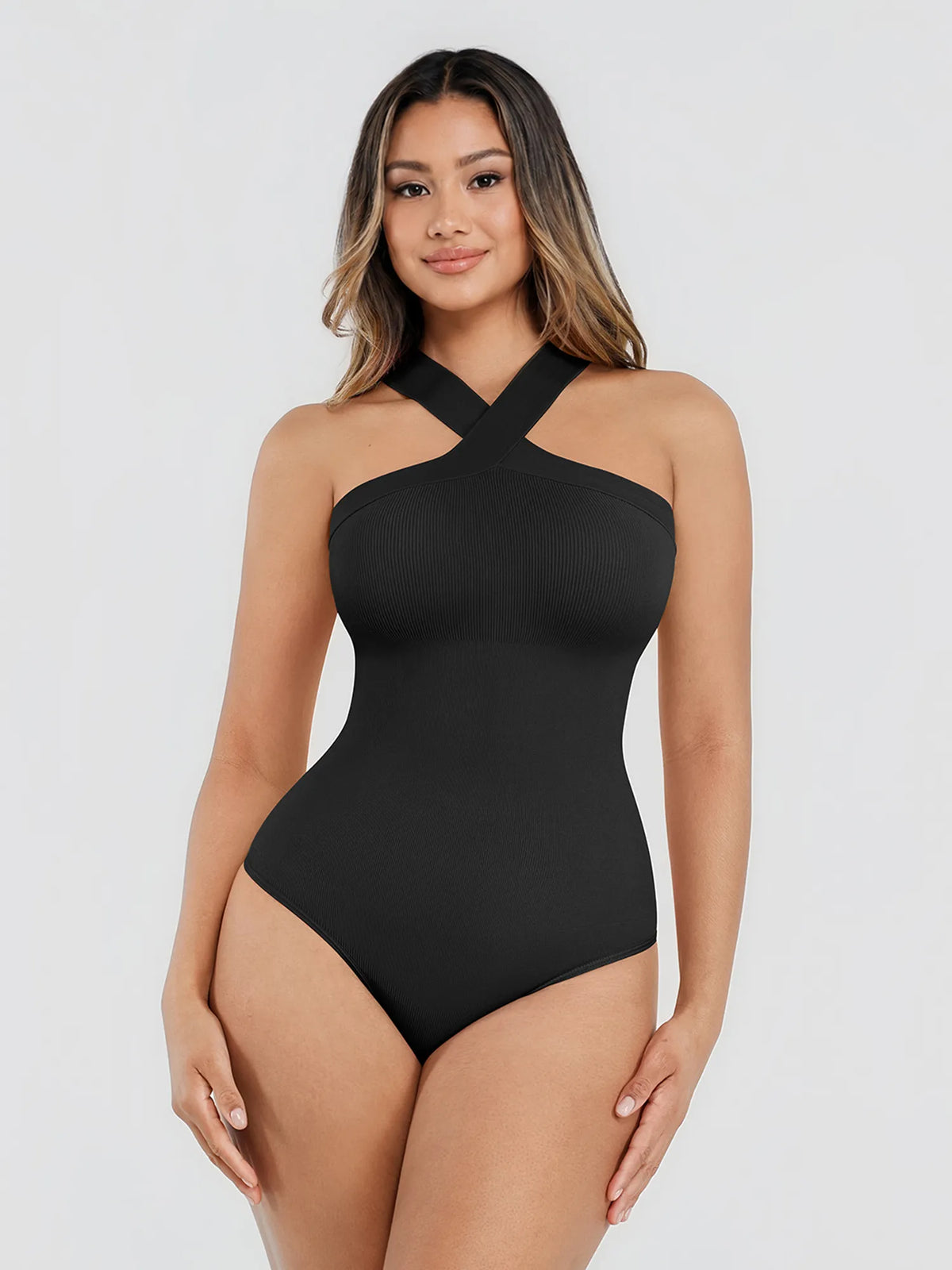 A woman modeling a black one-piece swimsuit with a halter-style crisscross neckline, photographed against a plain light gray studio background.