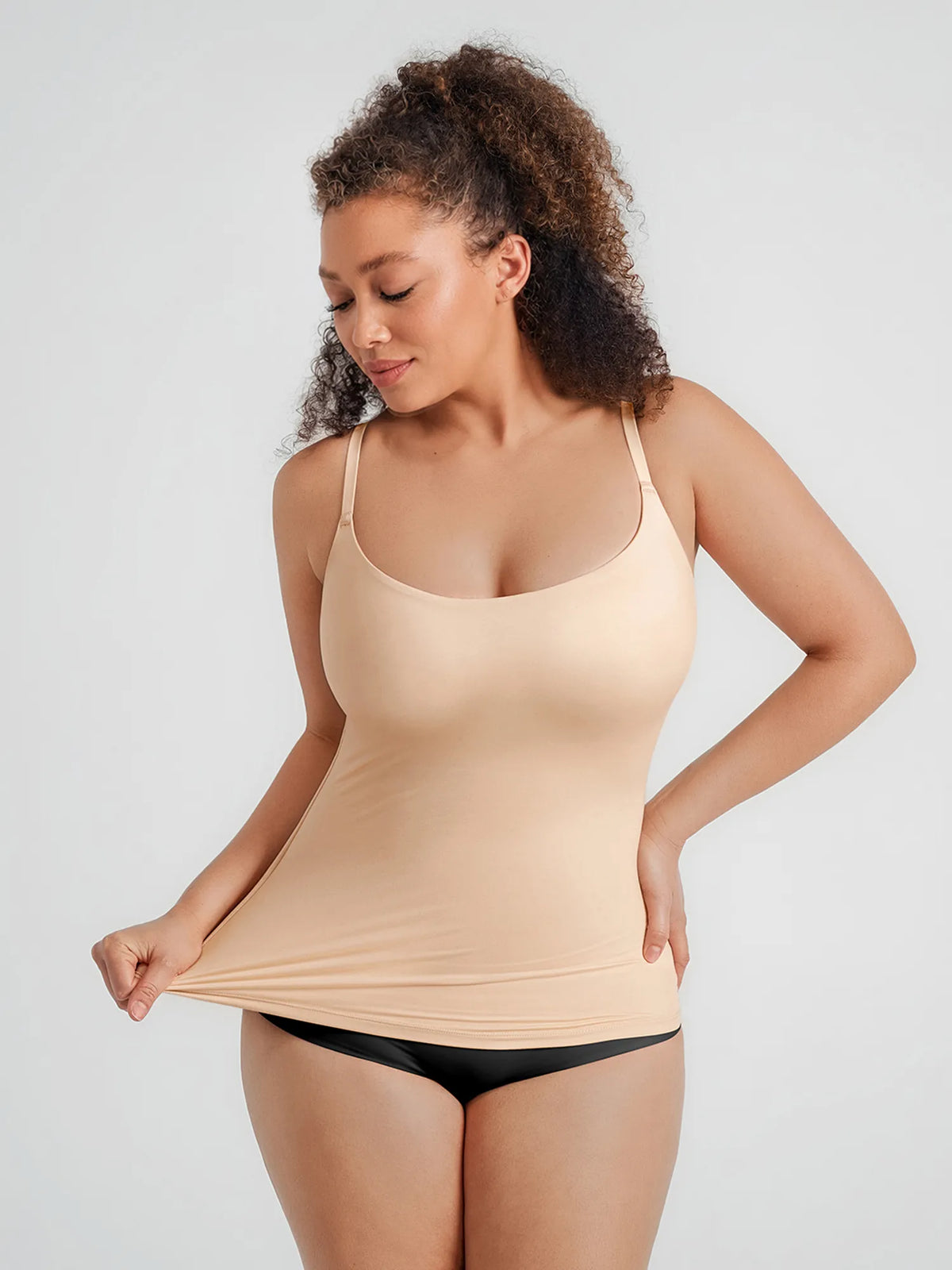 Front view of a model pulling the hem of a beige scoop-neck shaping cami top made with buttery-soft fabric and adjustable straps